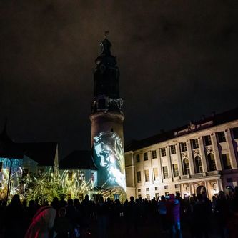 Die Fassade der Bastille, Turm und Schloss sind in der Nacht mit Szenen angestahlt, davor steht eine große Menschenmenge und betrachtet die Projektion