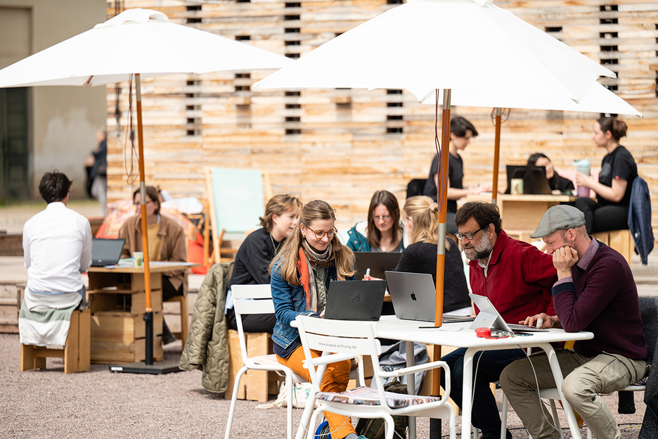 An mehreren Tischen vor dem Co-Labor arbeiten Personen an Laptops.