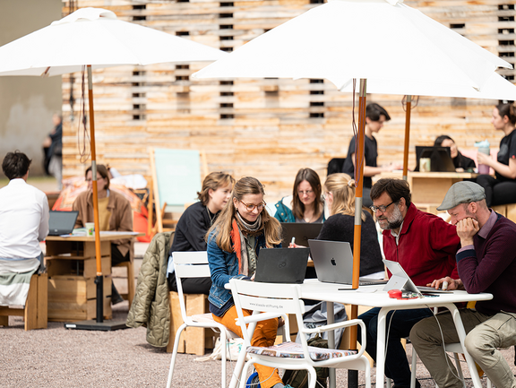 An mehreren Tischen vor dem Co-Labor arbeiten Personen an Laptops.