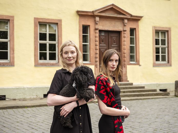 Zwei Personen mit einem schwarzen Pudel vor einem gelben Haus