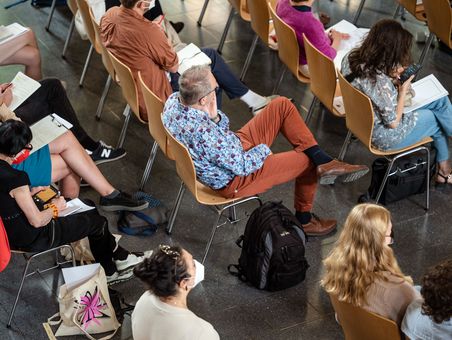 von oben fotografiert: Teilnehmende einer Tagung sitzen in Reihen auf Stühlen. Sie hören zu.