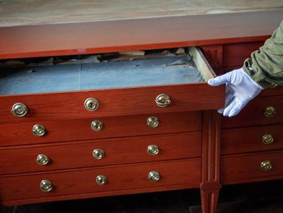 Eine behandschuhte Hand zieht eine Schublade aus einem historischen rotbraun leuchtenden Holzunterschrank.