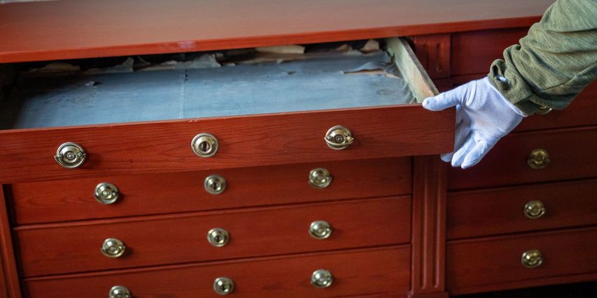 Eine behandschuhte Hand zieht eine Schublade aus einem historischen rotbraun leuchtenden Holzunterschrank.