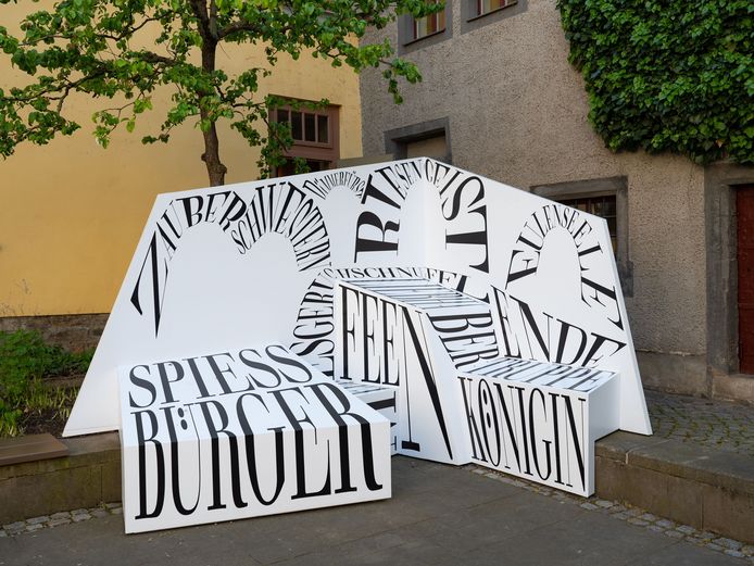 Eine Fotowand mit verschiedenen Wortschöpfungen, auf die sich gesetzt werden und vor der posiert werden kann, im Hintergrund eine Häuserecke und ein Baum