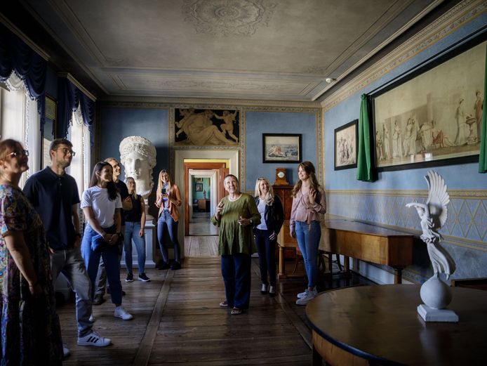 In a blue room with a view through a doorway, several people stand around a guide and listen to what they have to say.