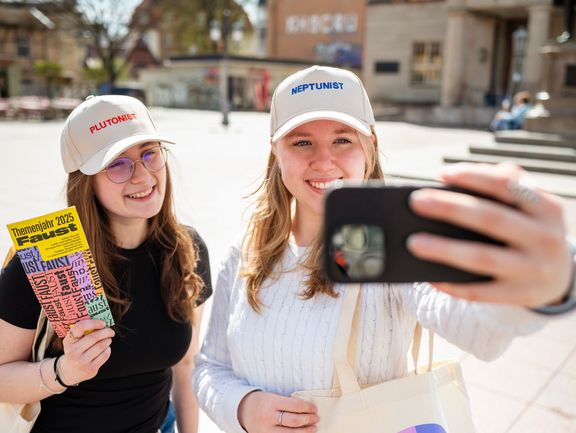 Zwei personen tragen Basecaps mit den Aufdrucken "Plutonist" und "Neptunist" und machen ein Selfie.