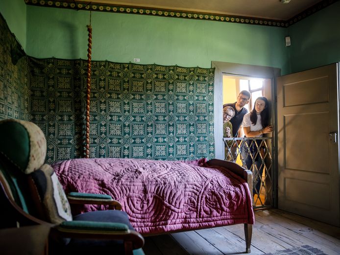 Drei Leute spähen über den Türrahmen in einen mit Wandteppich dekorierten Raum, in dem ein Bett mit Überzug steht