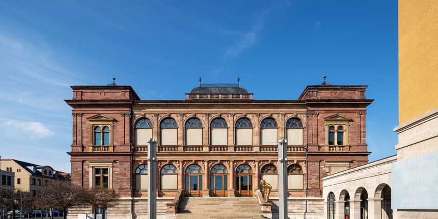Vom Vorplatz aus wird die symmetrische Vorderansicht des rot-bräunlich-backsteinfarbenen Museums mit seinem Treppenaufgang hin zum oberen Eingangsbereich gezeigt.
