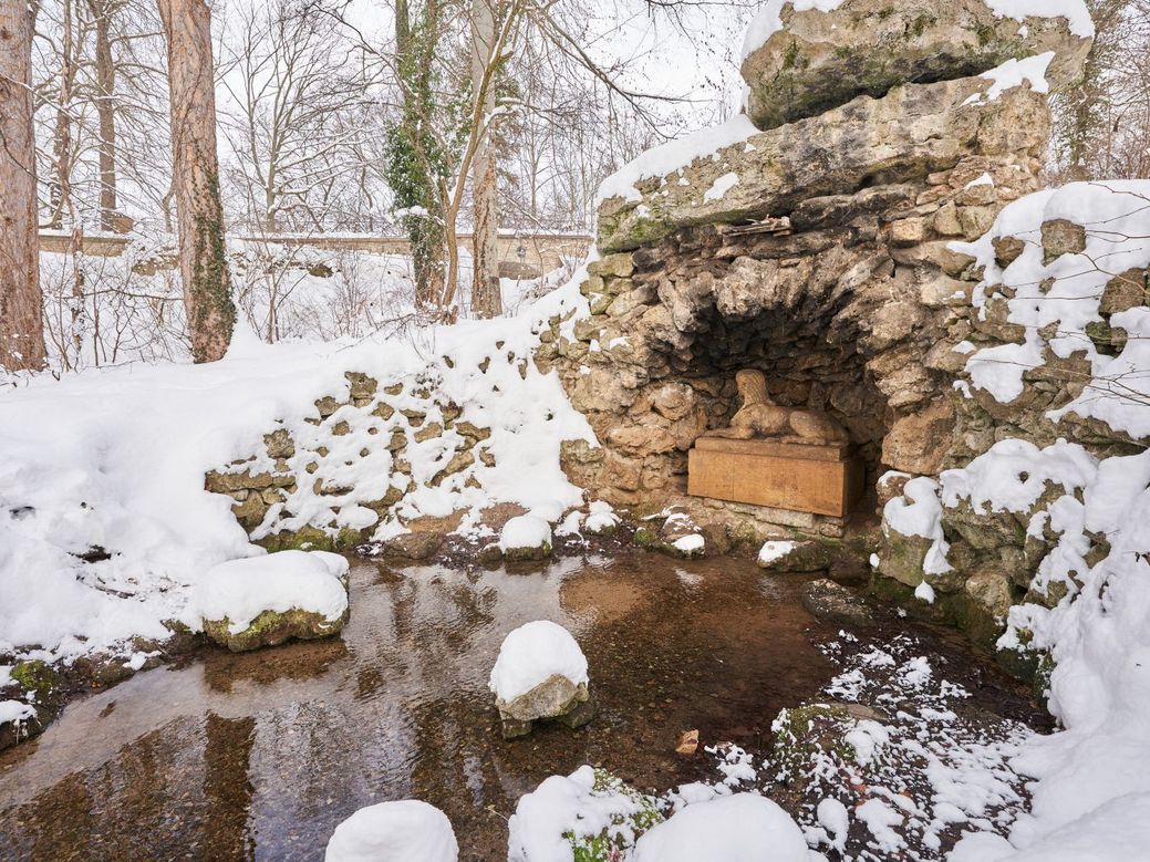 Im Vordergrund einer Schneelandschaft befindet sich eine Sphinx unter einer kleinen Felshöhle. Unter ihr befindet sich eine Flussquelle. 