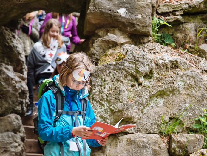 Mädchen mit Rucksack blättert durch rotes Rucksacktour-Heft