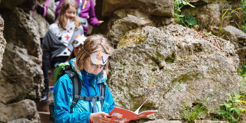 Mädchen mit Rucksack blättert durch rotes Rucksacktour-Heft