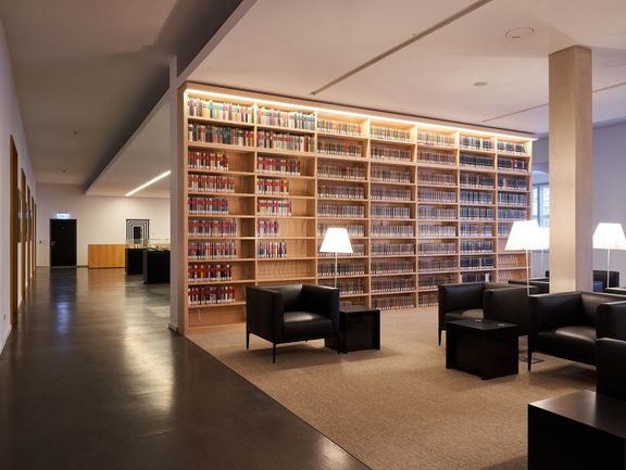 Die schwarzen Sessel der Leselounge stehen vor einem großen, hell beleuchteten Bücherregal, das mit zahlreichen Büchern gefüllt ist. Der Raum ist hell und einladend gestaltet, mit einem Teppichboden und modernen Leuchten an der Decke. Ein Gang führt zu weiteren Bereichen des Gebäudes.