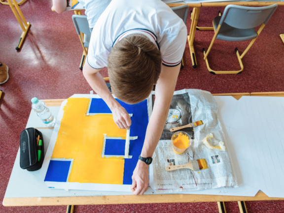 Draufsicht auf ein Kind, das auf Papier mit Farbe und Klebestreifen Quadrate gestaltet.