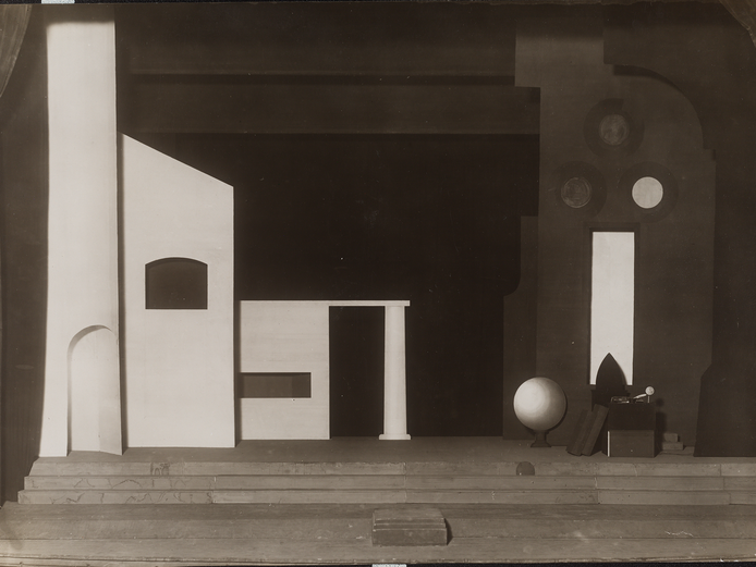 Das schwarz-weiße Bild zeigt eine abstrakte Bühnenbild-Szene. Im Vordergrund sind mehrere geometrische Formen und Strukturen zu sehen, darunter rechteckige und runde Öffnungen sowie eine Kugel. Die Szene ist in starken Kontrasten von Licht und Schatten gehalten, was eine dramatische Atmosphäre erzeugt. Im Hintergrund sind weitere abstrakte Elemente und Formen zu erkennen.