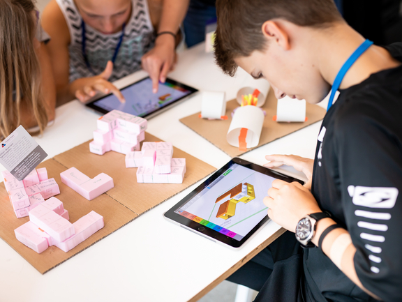 Jugendliche beugen sich über ein Architekturmodell und ein Junge erarbeitet an einem Tablet ein digitales Modell für den Bau.