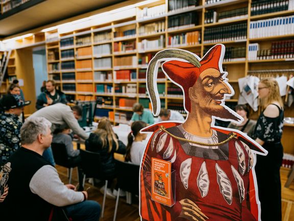 Im Vordergrund steht ein Mephisto-Pappaufsteller, im Hintergrund sitzen Personen an einem langen Tisch.