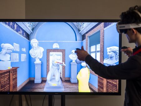 Goethe-Live-3D Eine Person mit einem Virtual-Reality-Headset und Controllern in den Händen steht vor einem großen Bildschirm. Auf dem Bildschirm ist eine virtuelle Szene zu sehen, die wie ein Museumsraum aussieht. Der Raum hat blaue Wände und ist mit verschiedenen Statuen und Büsten auf Sockeln dekoriert. Die Person interagiert mit der virtuellen Umgebung, indem sie einen der Controller in Richtung des Bildschirms bewegt. Im Hintergrund der virtuellen Szene ist ein Fenster zu sehen, durch das Tageslicht hereinfällt.