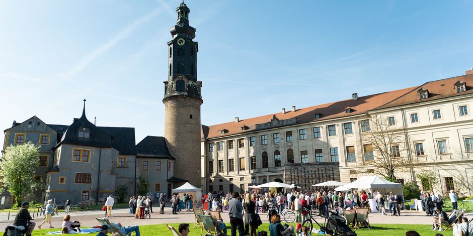 Menschen sitzen und stehen auf einer grünen Wiese. Im Hintergrund ist ein großes Schloss.