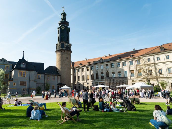 Menschen sitzen und stehen auf einer grünen Wiese. Im Hintergrund ist ein großes Schloss.