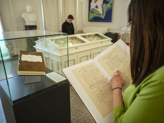 Ein altes Buch in einer Vitrine und eine Person, die in einem anderen Heft blättert