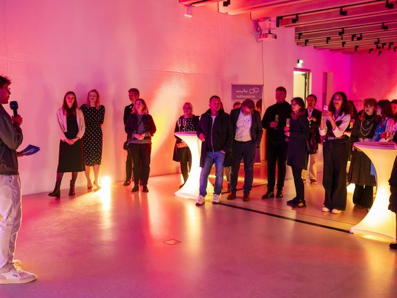 Links im Bild steht ein Mann und spricht mit einem Handmikrofon zur Menschengruppe rechts im Bild.