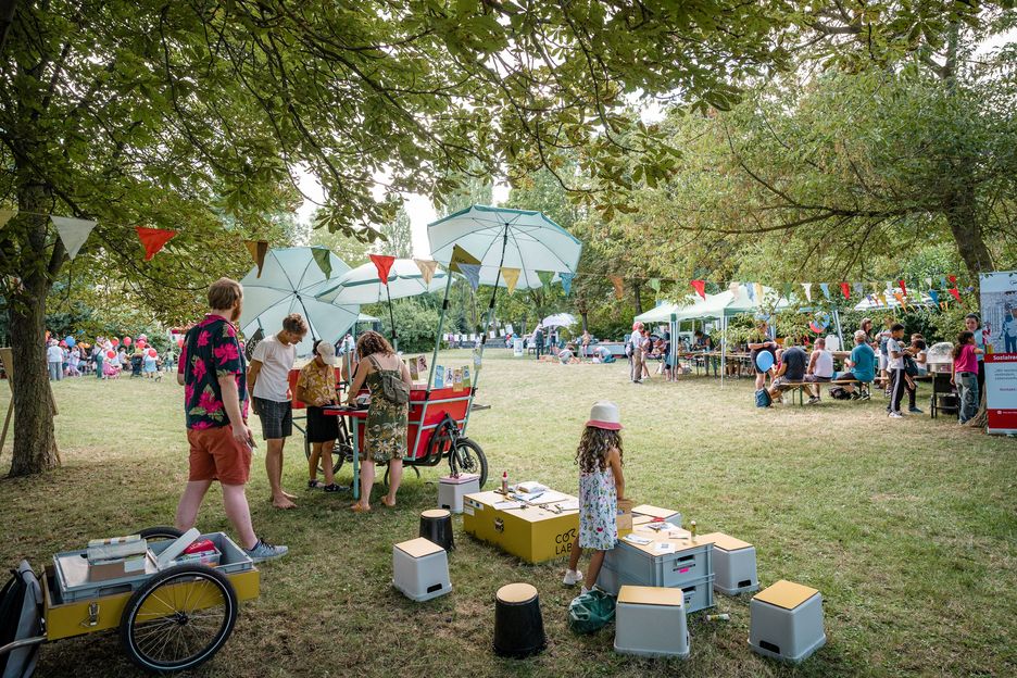 Auf einer sommerlichen Wiese sind mehrere Zelte aufgebaut. Daneben steht ein Lastenrat mit Sonnenschirmen und Spielkisten. Dies ist das Co-Labor unterwegs.
