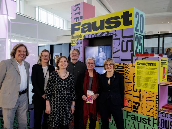 Sechs Personen stehen vor einer Wand, auf der "FAUST" zu lesen ist.